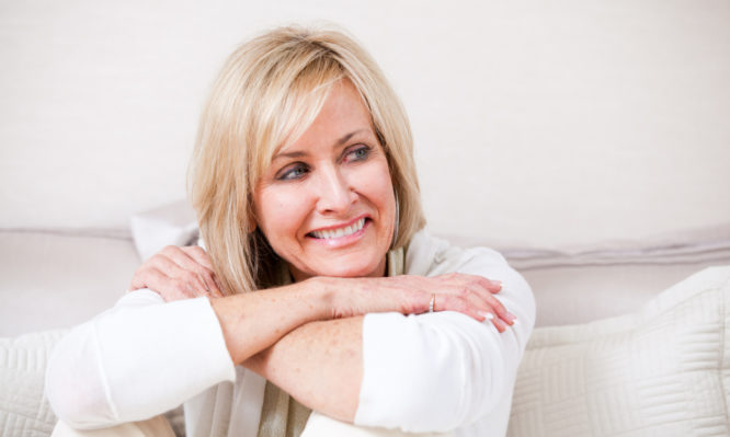 Happy mature woman in her 50s. She is sitting with her arms crossed. Woman is resting her chin on her crossed arms. She is wearing an off white color with the similar off white color in the background. She is smiling and looking of to the side.