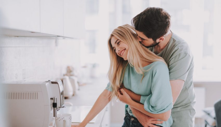 Bearded guy standing behind attractive blonde girl and kissing her. They are waiting for hot drink