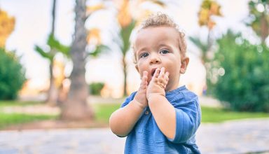 Sad little boy putting fingers on mouth touching gums because toothache at the park on a sunny day. Beautiful blonde hair male toddler in pain for new baby teeth outdoors