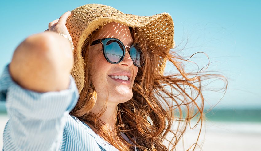 Side view of beautiful mature woman wearing sunglasses enjoying at beach. Young smiling woman on vacation looking away while enjoying sea breeze wearing straw hat. Closeup portrait of attractive girl relaxing at sea.